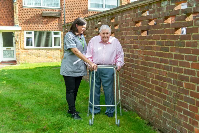 Carer supporting elderly man with a walking frame in the garden